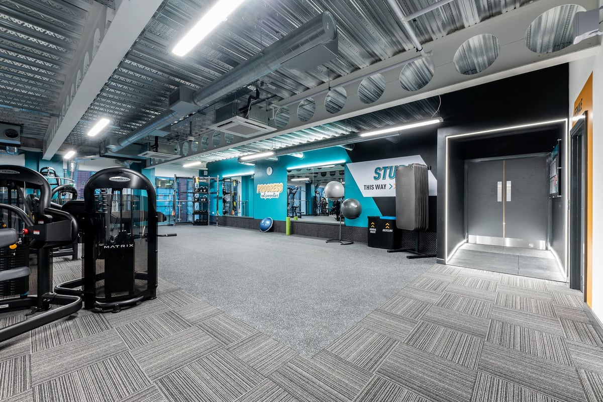 PureGym Manchester First Street: Fitness studio with weight machines, turf floors, entry gates, and teal accents.