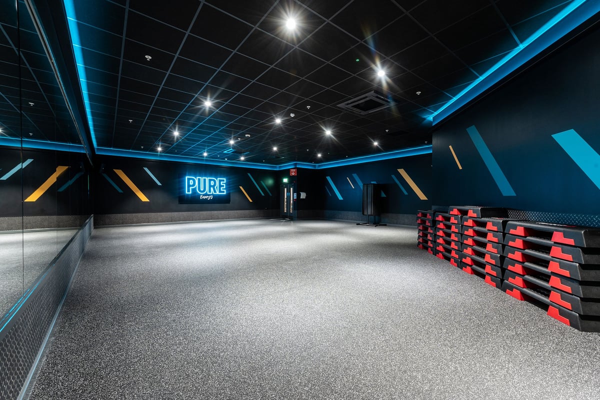 PureGym Bracknell: Black mirrored studio with patterned carpet, angled lights, freeweight bars, and lineup of weighted sleds.
