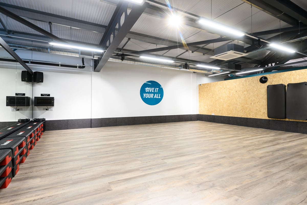 PureGym Barnstaple: Large open studio with wall mirrors, rubberized flooring, weight racks, and speakers mounted overhead.
