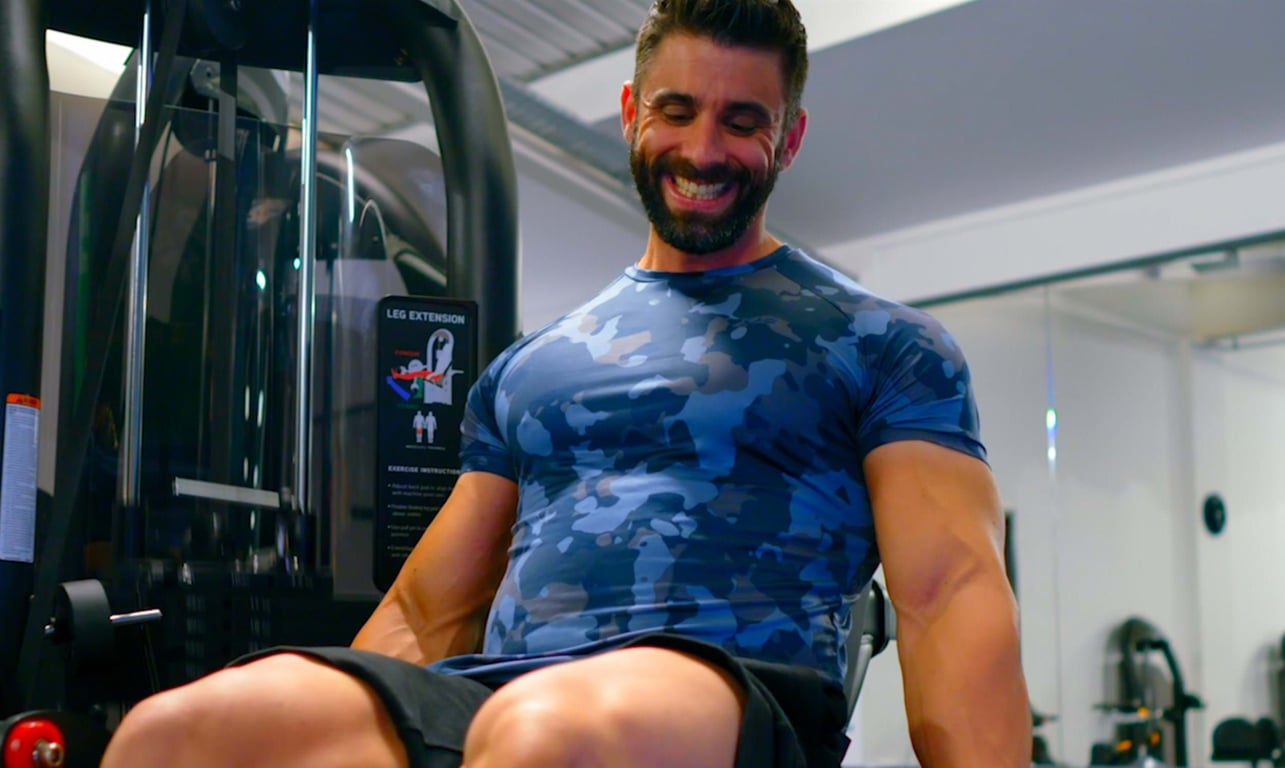 Man in blue camo shirt smiling while using leg extension machine at gym