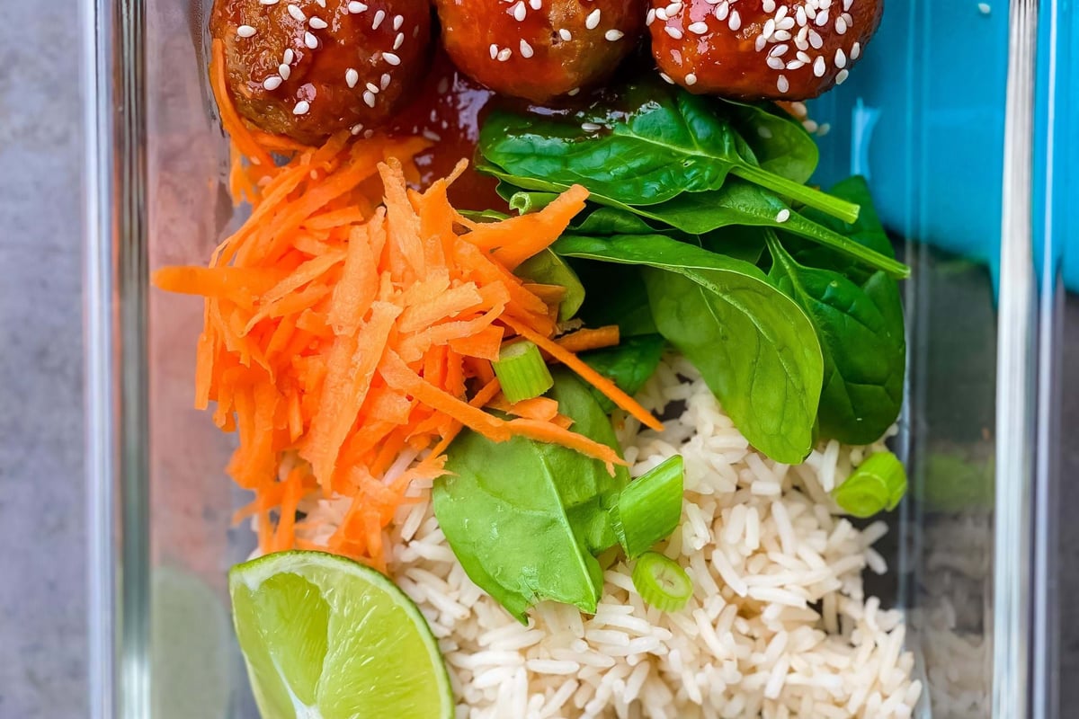 Three meatballs with sauce and sesame seeds, shredded carrot, spinach, rice, and lime wedg