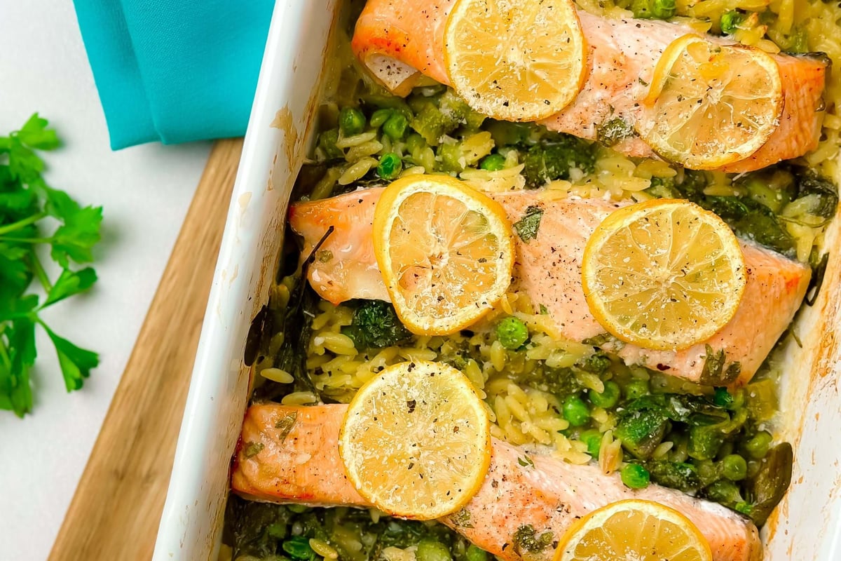 Baked salmon fillets topped with lemon slices on a bed of rice and peas in a white dish