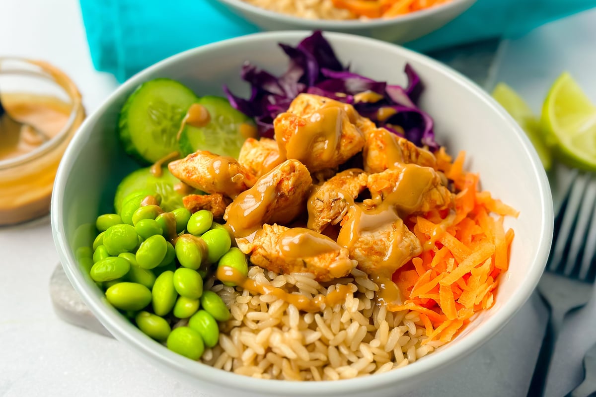 Bowl of chicken with brown rice, edamame, cucumber, red cabbage, carrot and lime wedge