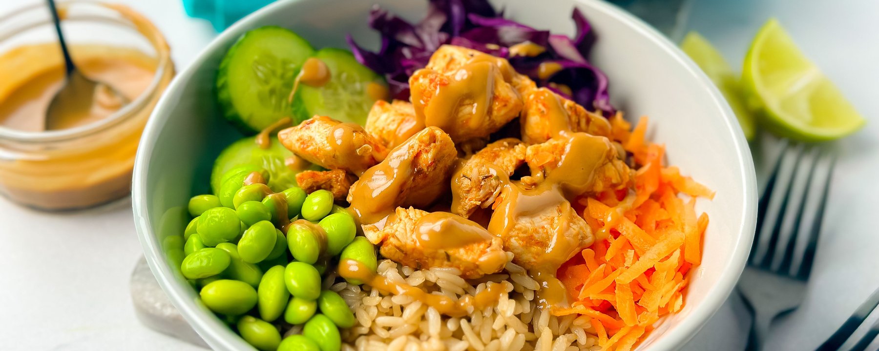 Bowl of chicken with brown rice, edamame, cucumber, red cabbage, carrot and lime wedge