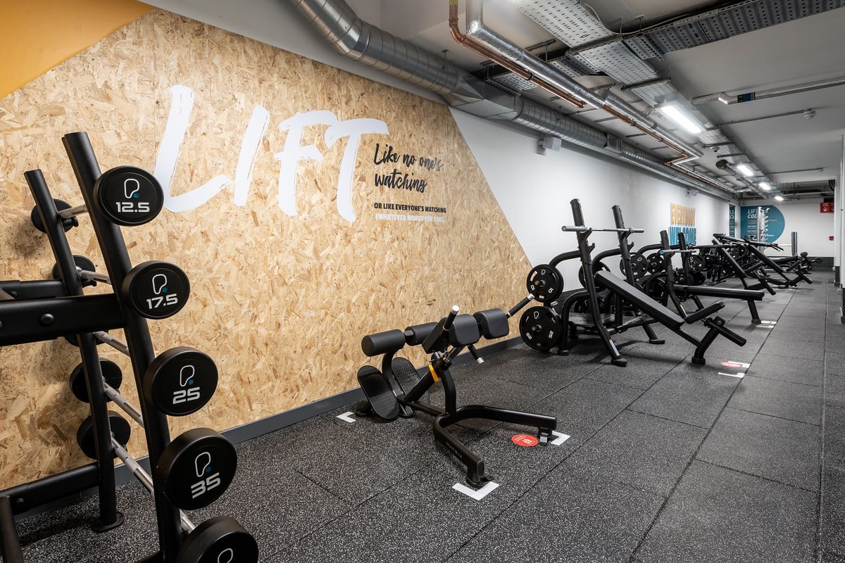 PureGym London Holloway Road: Rows of weight benches, barbells, weight plates, and a motivational wall mural inside a gym.