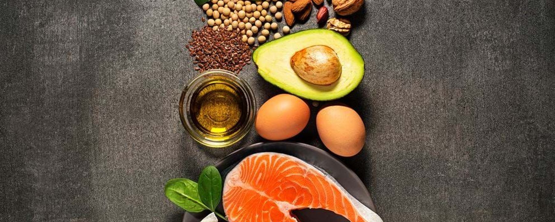 Salmon steak, avocado, eggs, nuts, seeds, spinach leaves, and oil on dark surface.