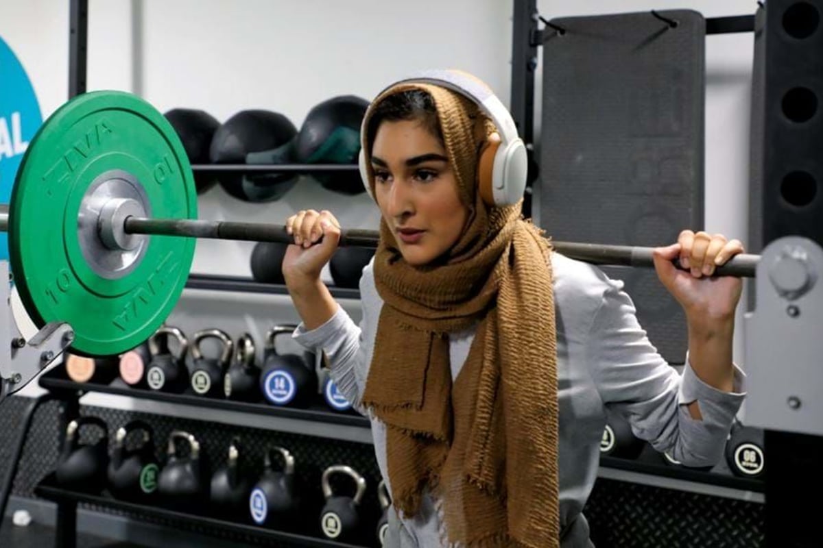 Woman wearing hijab and headphones lifting a barbell in a gym with kettlebells behind her