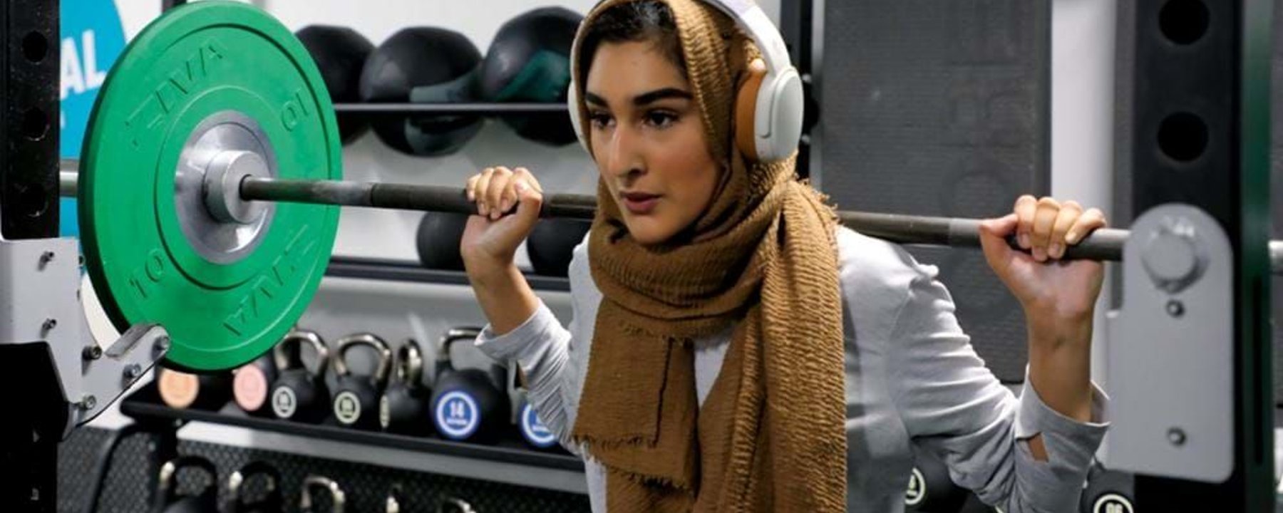Woman wearing hijab and headphones lifting a barbell in a gym with kettlebells behind her