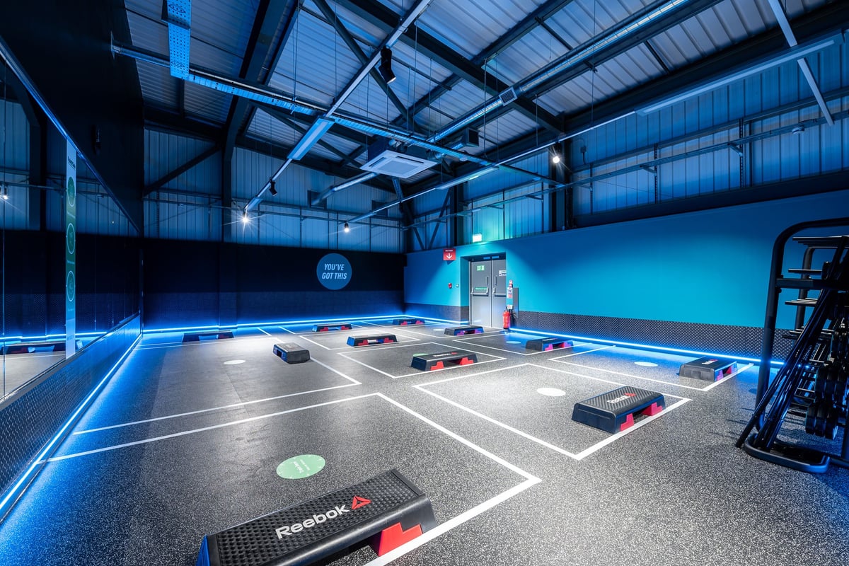 PureGym Birmingham Longbridge: Indoor gym studio with rubberized flooring, exercise equipment, and colored accent lighting.