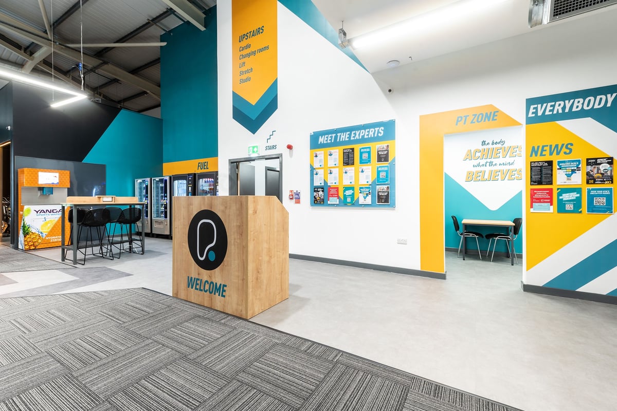 PureGym Macclesfield Silk Road: Checkout area with colorful signage, tables and chairs for consultations.
