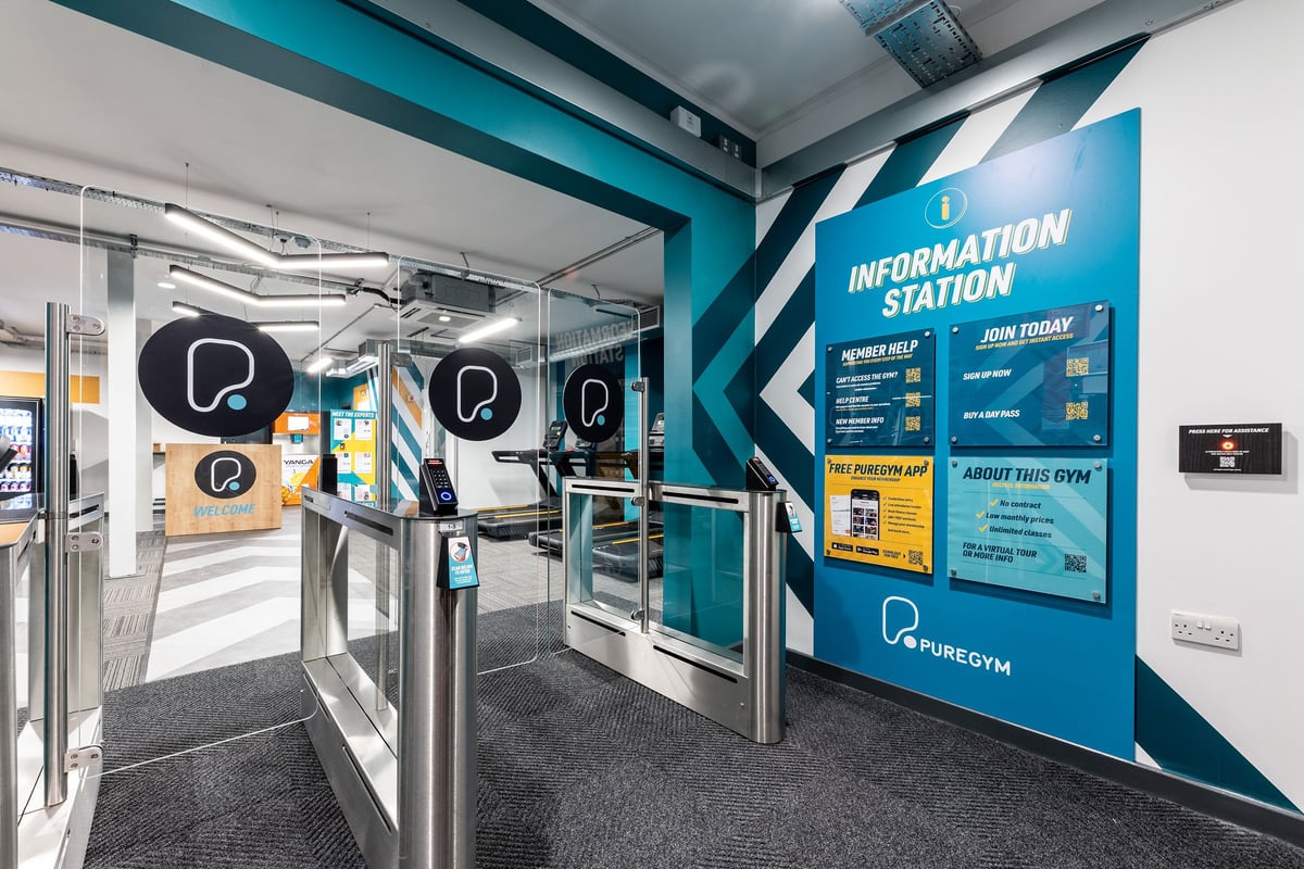 PureGym Manchester First Street: Information station showing directional signage, turnstiles, teal branding.