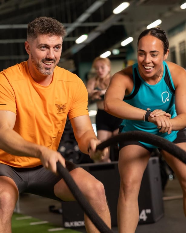 Zwei lächelnde Personen trainieren zusammen mit Battle Ropes in einer hellen PureGym-Anlage