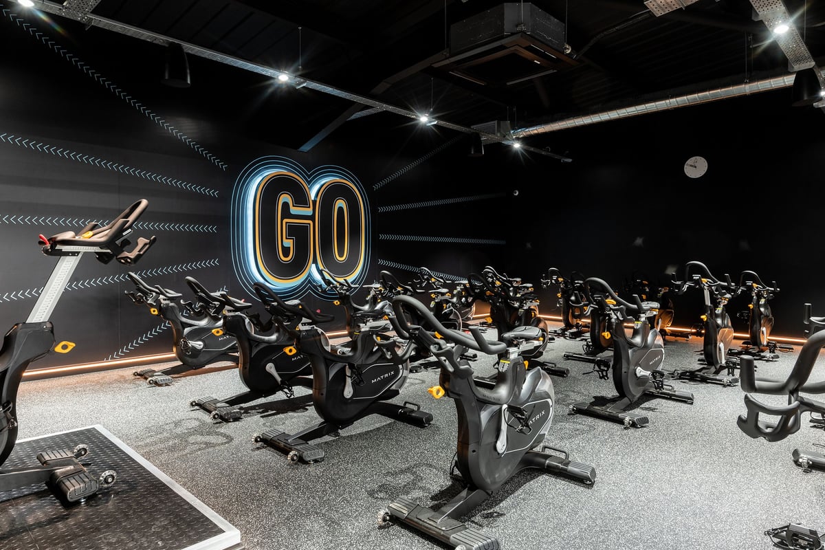 PureGym Cardiff Western Avenue: Rows of stationary bikes, neon 'GO' sign, mirrored walls in a dimly lit studio.