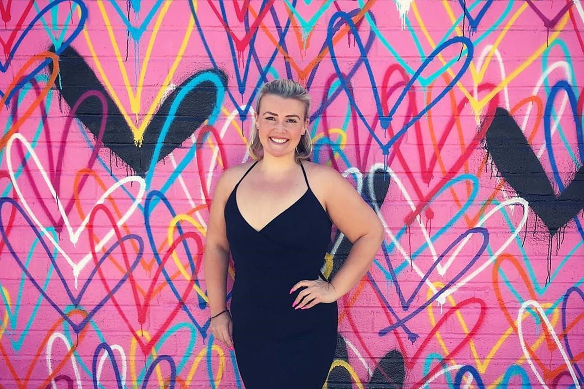 Woman in black dress smiling in front of a colourful heart graffiti wall