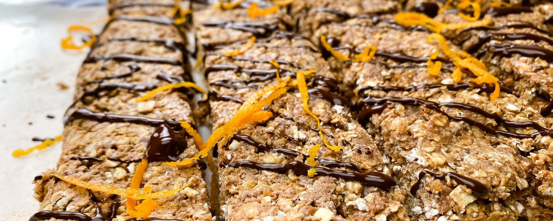Oat-based protein flapjacks drizzled with dark chocolate and orange zest on white surface