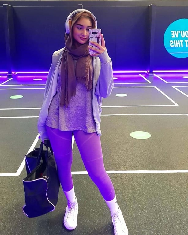 Woman in purple leggings and brown scarf taking a mirror selfie in a gym with neon lights