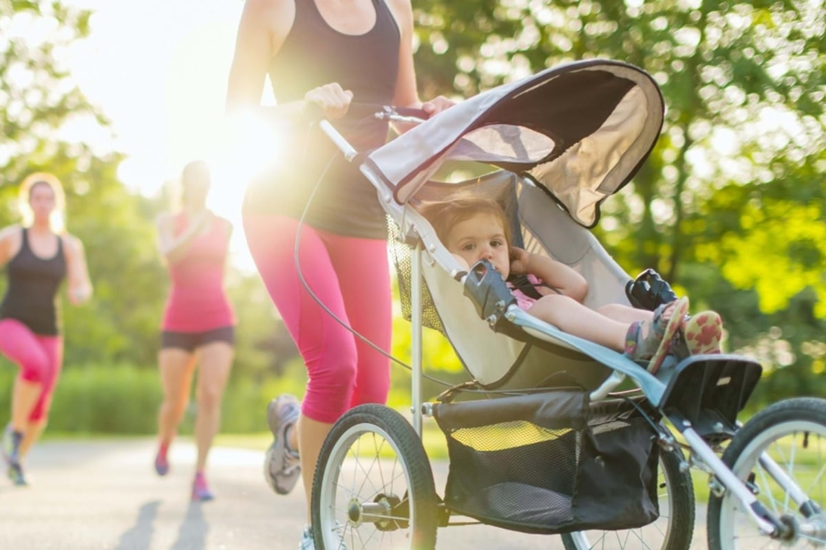 Woman jogging with a child in a stroller on a sunlit park path with others running behind