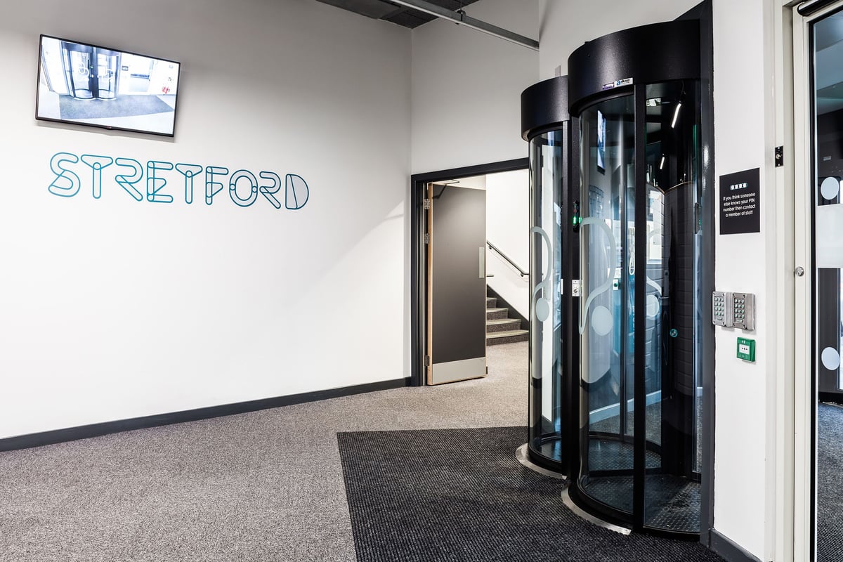 PureGym Manchester Stretford: Glass revolving door stall and stretford sign on wall indicating fitness studio entrance.
