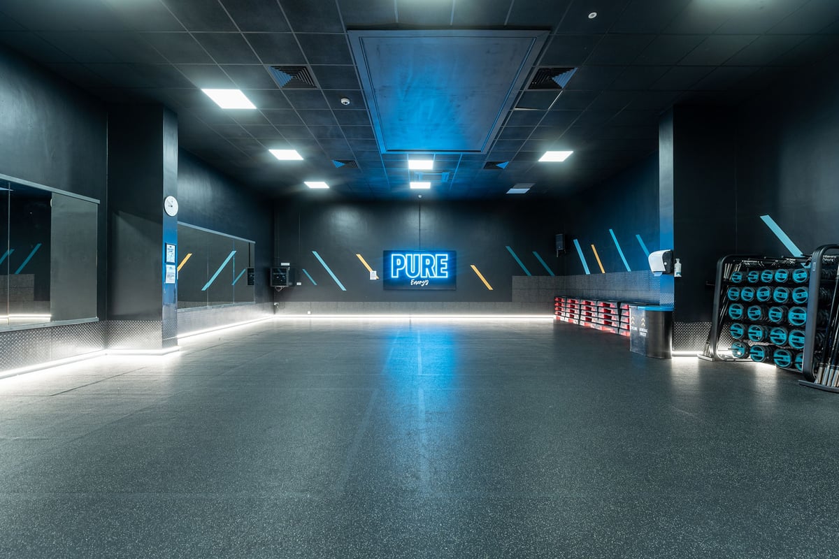 PureGym Northolt: Large open studio space with blue lighting accents, Pure branding, and storage racks for equipment.