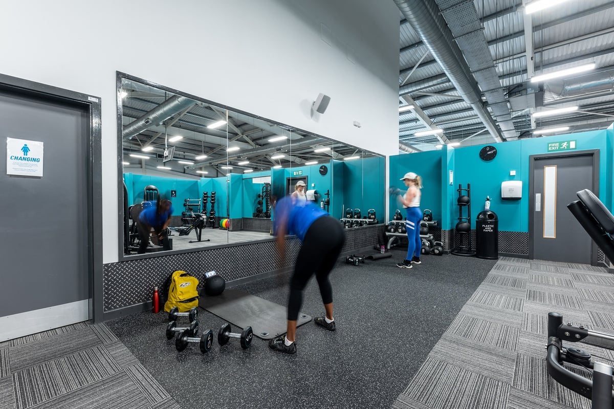PureGym Birmingham West: Gym studio with weight benches, free weights, rubberized flooring, and teal accent walls.