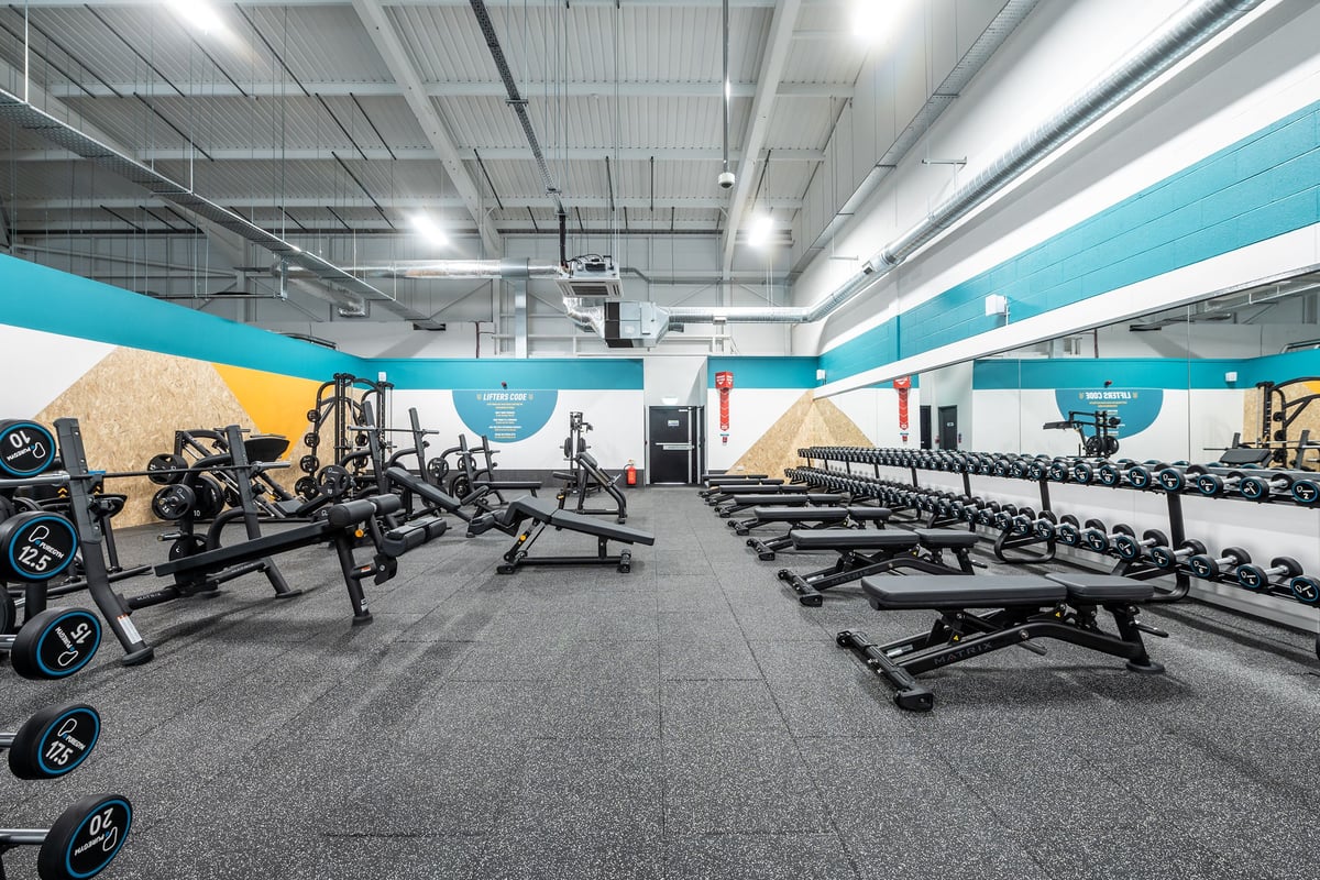 PureGym Winchester: Weightlifting area with benches, racks, free weights, and mirrored walls in a modern gym interior.
