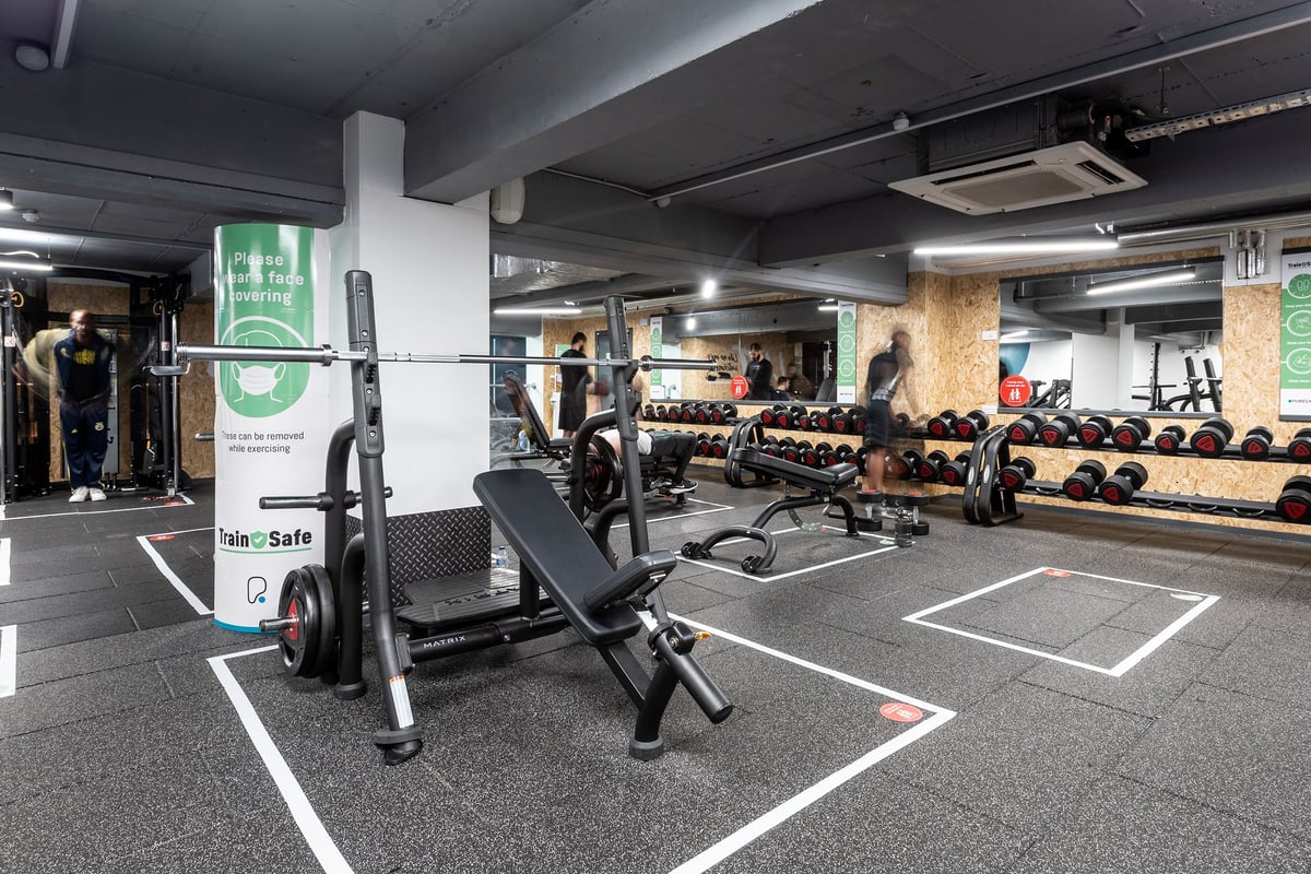 PureGym London Sydenham: Weight benches, squat racks, dumbbell racks, rubberized floor markings in a basement gym space.