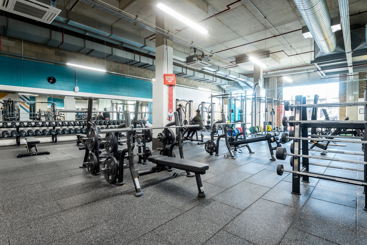 PureGym London Woolwich: Weight benches, dumbbells, racks of free weights, and rubberized flooring in a well-lit gym space.
