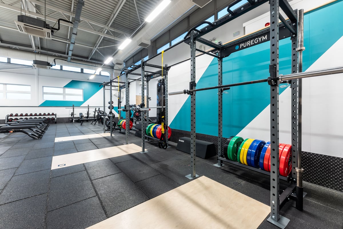 PureGym London Edgware: Squat racks, weight plates, rubberized flooring in a gym with teal accent walls and storage racks.