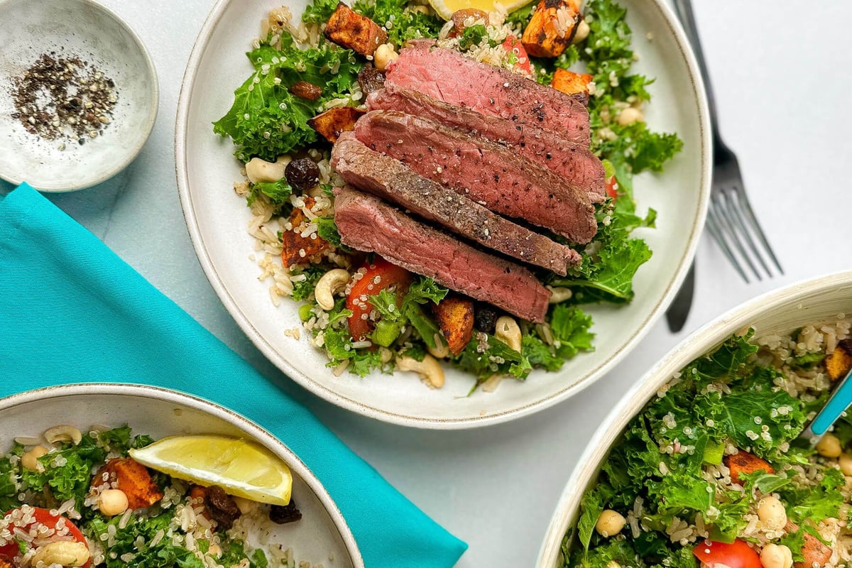 Sliced steak on a bed of kale, quinoa, nuts, and roasted vegetables with lemon wedges.