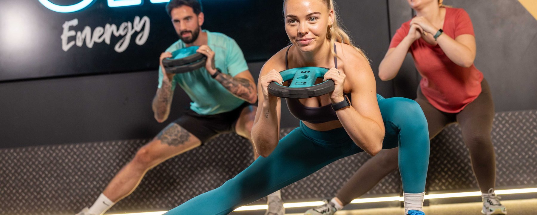 Three people stretching with weights in a gym with a PURE Energy sign behind them