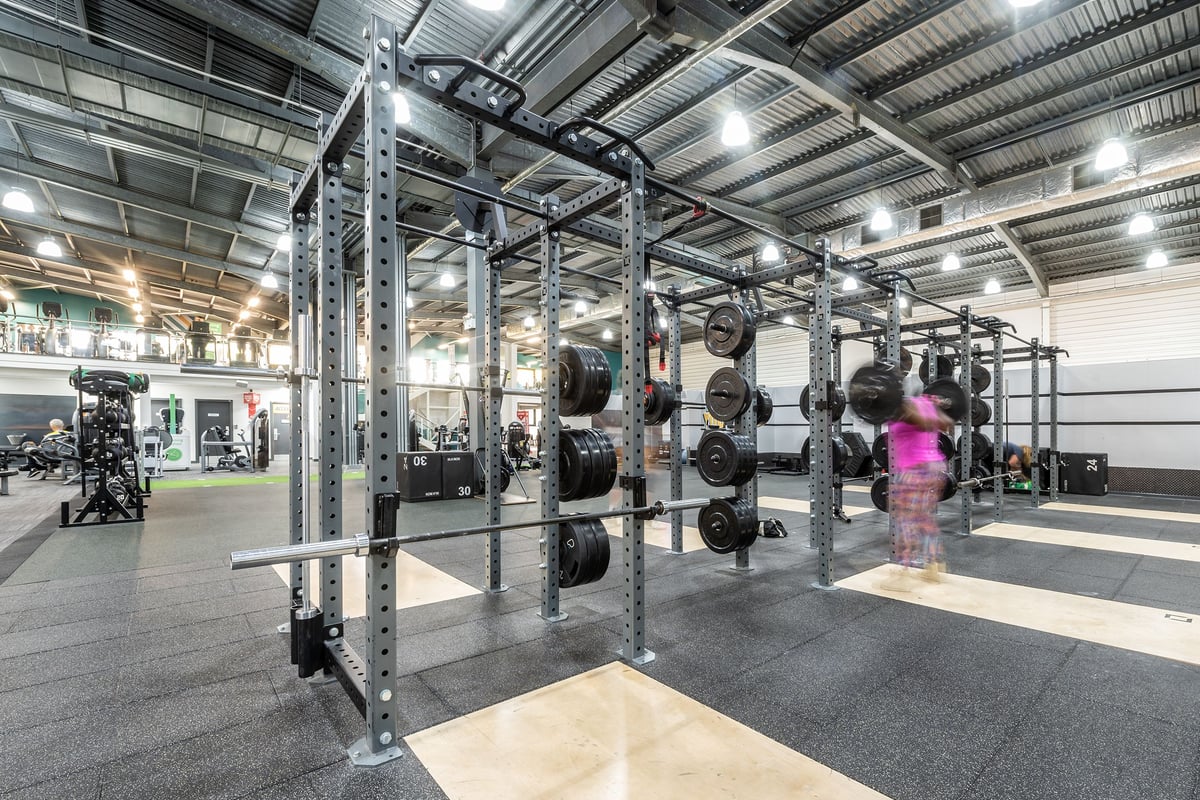 PureGym Milton Keynes Winterhill: Squat racks with weight plates on rubberized flooring in an industrial-style gym interior.