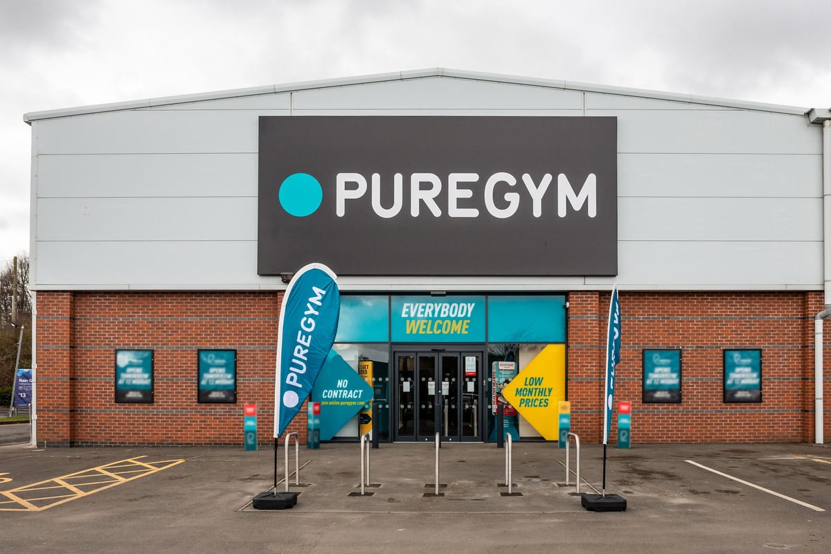 PureGym Market Harborough: Gym exterior with signage, flagpoles displaying brand, and parking spaces.