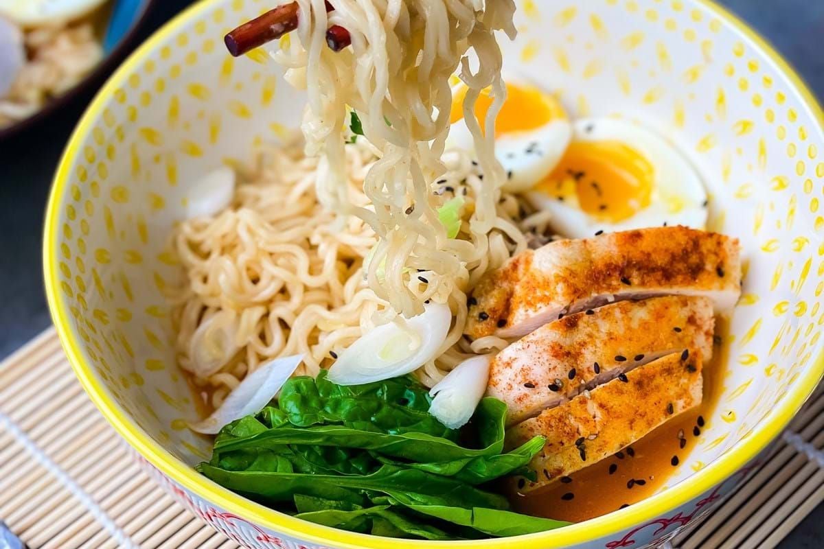 Chopsticks lifting noodles from a bowl with chicken, spinach, boiled egg, and broth.