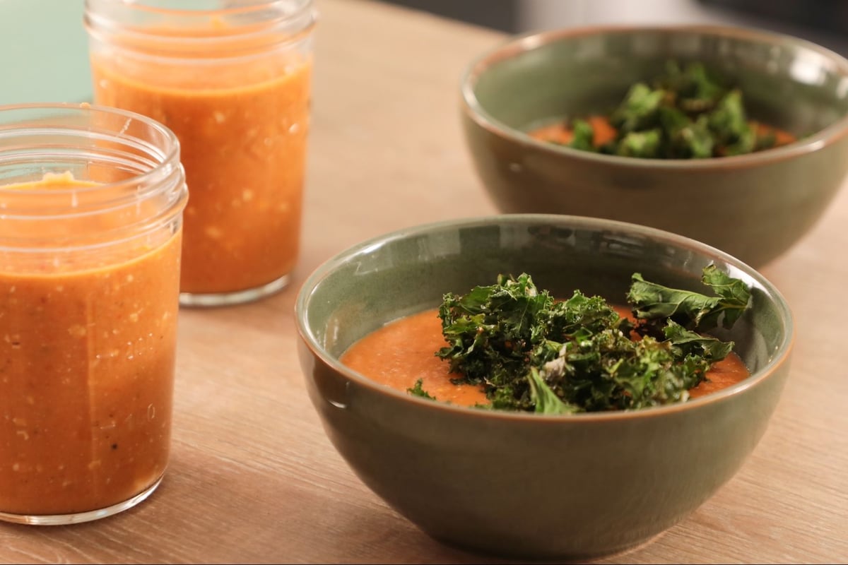 Two green bowls of soup with kale and two jars of orange soup on a wooden table