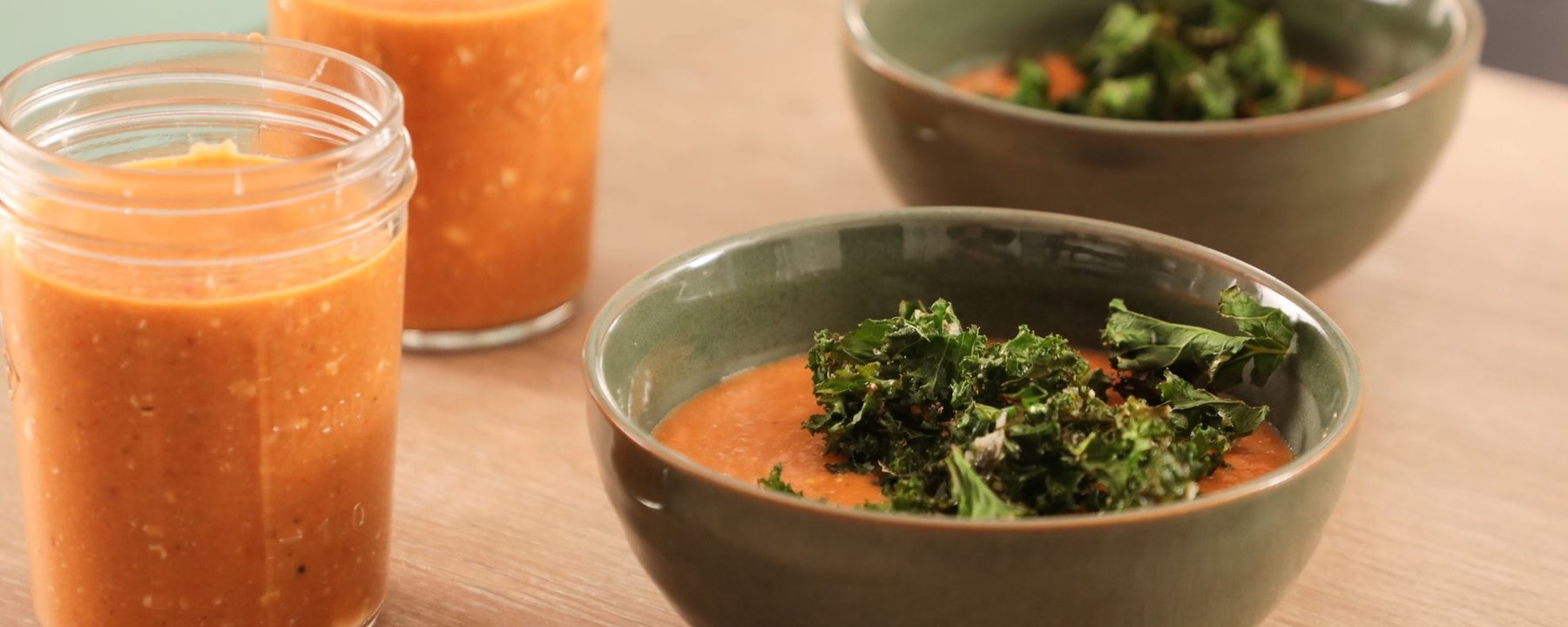 Two green bowls of soup with kale and two jars of orange soup on a wooden table