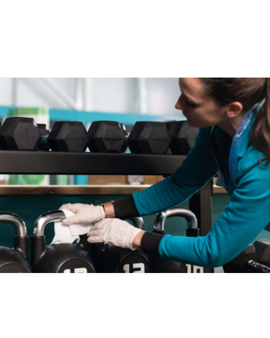 Woman in blue top cleaning dumbbells in a modern, bright gym studio with equipment racks