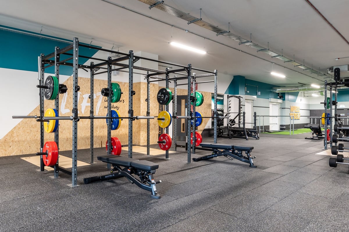 PureGym London Feltham: Rig racks, weight plates in multiple colours, benches, rubberized flooring in a gym setting.