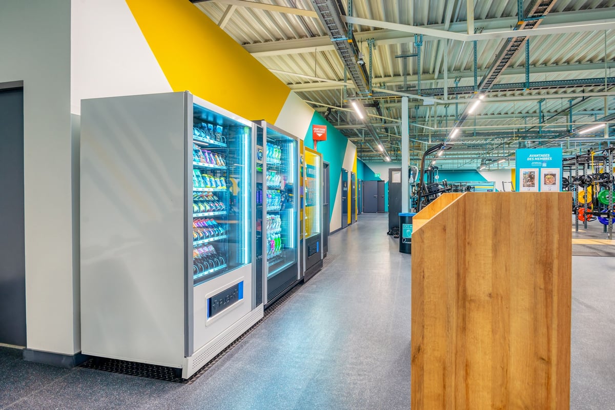 PureGym Martigny: Kühlautomaten säumen einen Korridor mit gelben Akzenten, Holzpaneelen und Metalldeckenbalken.