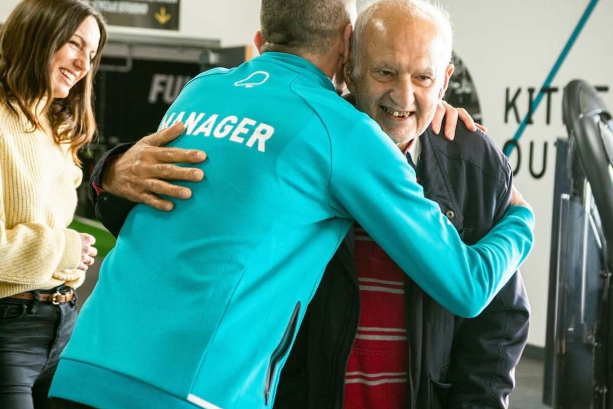 Man in blue jacket labelled manager hugging elderly man with walker while woman smiles nea
