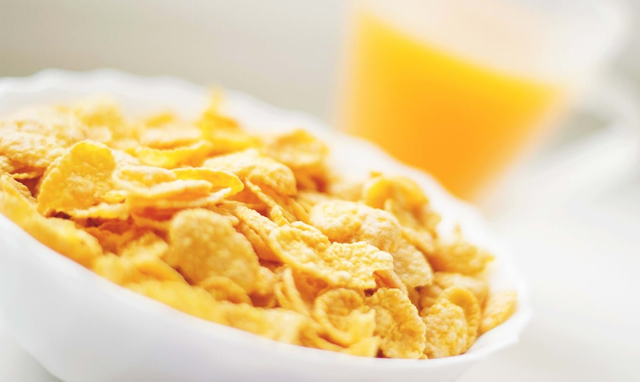 Cornflakes in a white bowl with a glass of orange juice in the background