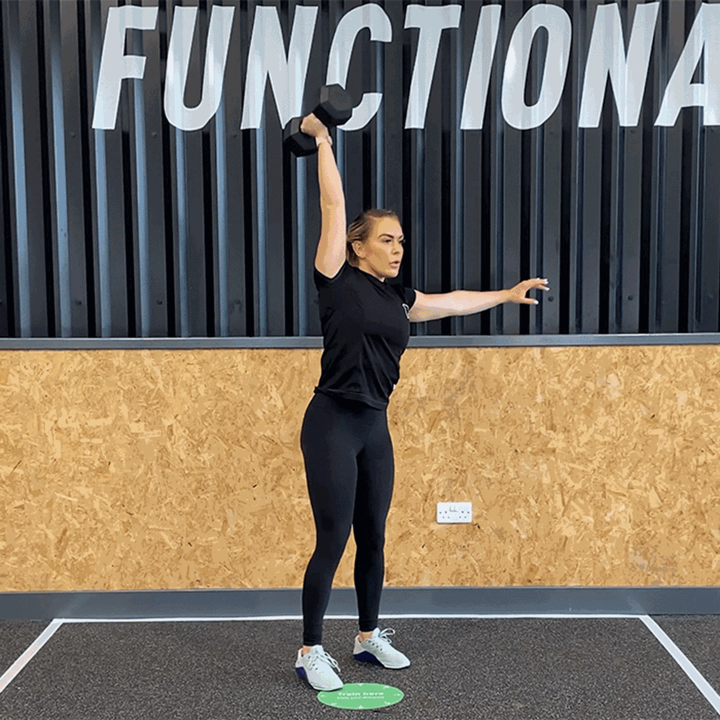 Woman lifting dumbbell overhead in gym with FUNCTIONAL sign behind her