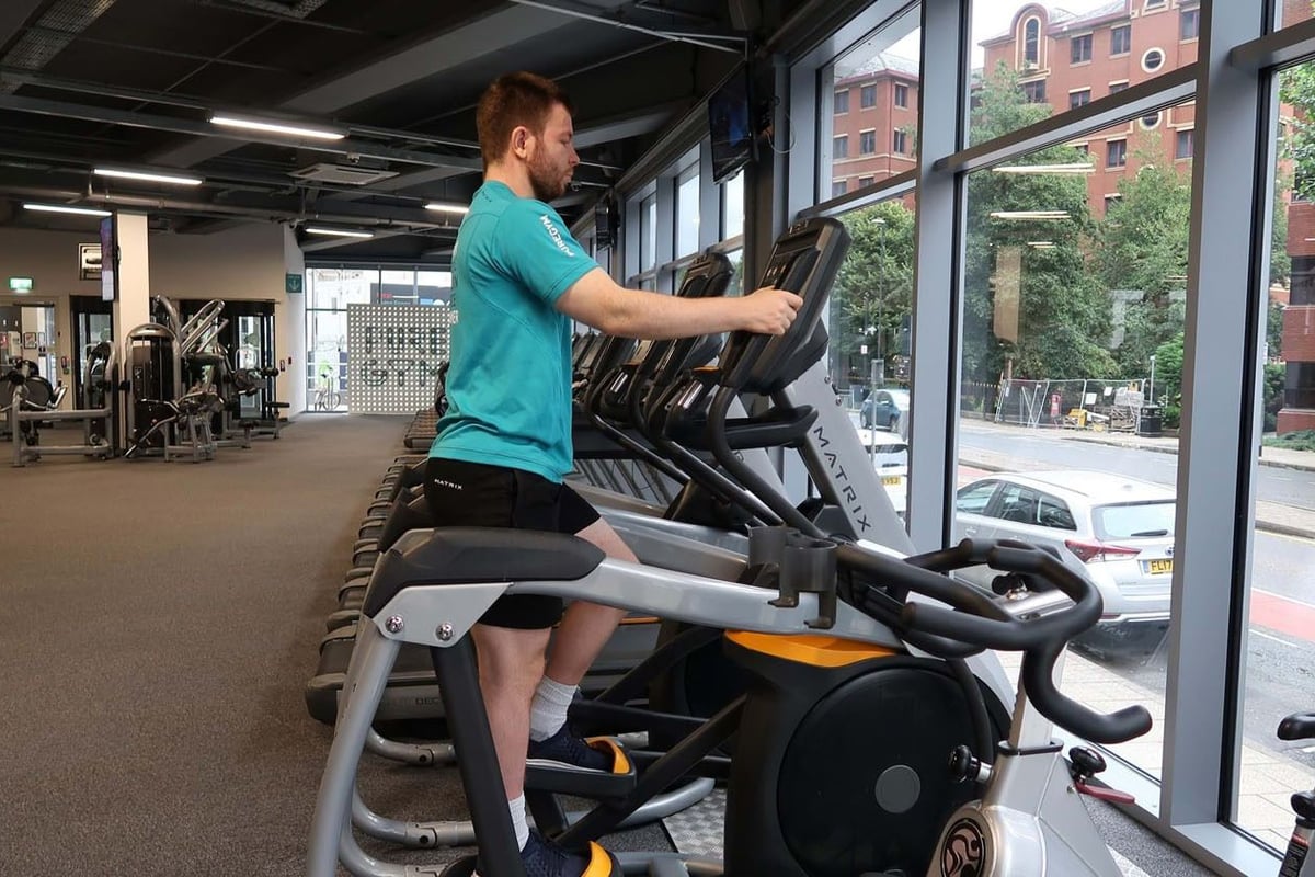 Man exercising on a stepper machine in a gym with large windows overlooking the street