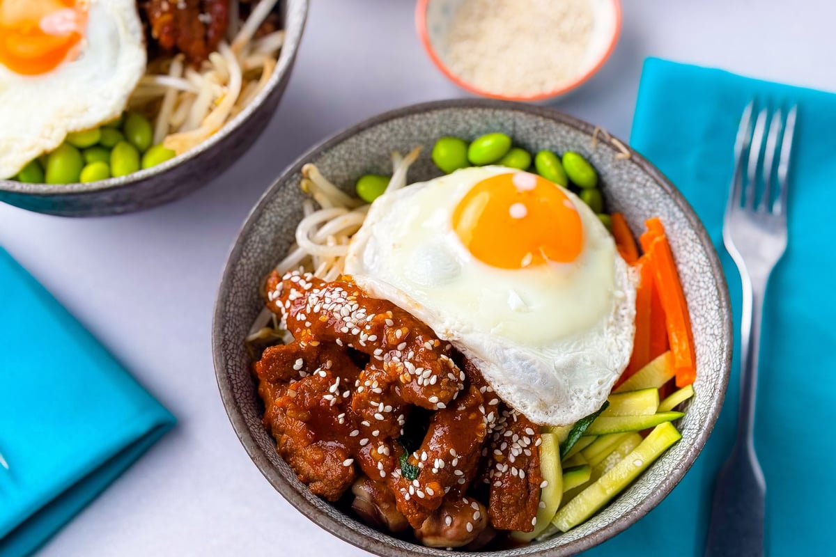 Bowl with fried egg, sesame chicken, edamame, carrots, and zucchini strips with fork