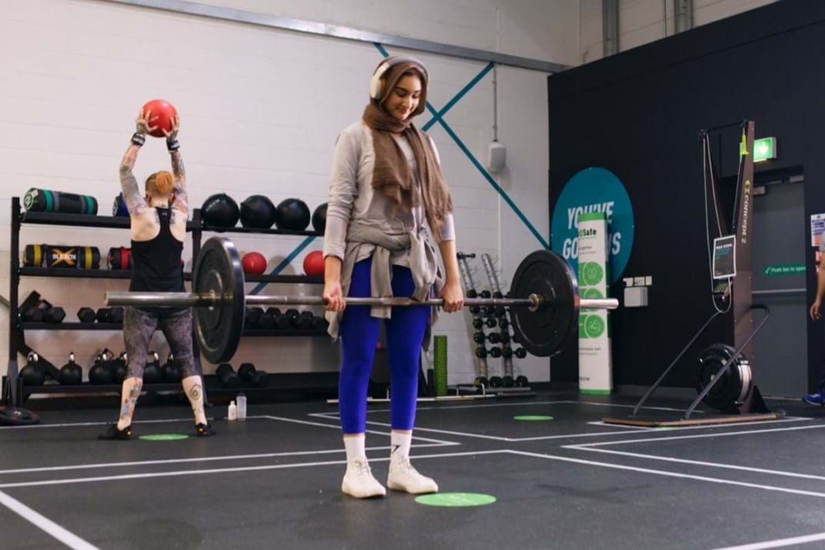 Woman in hijab lifting barbell while another person holds a red medicine ball in gym