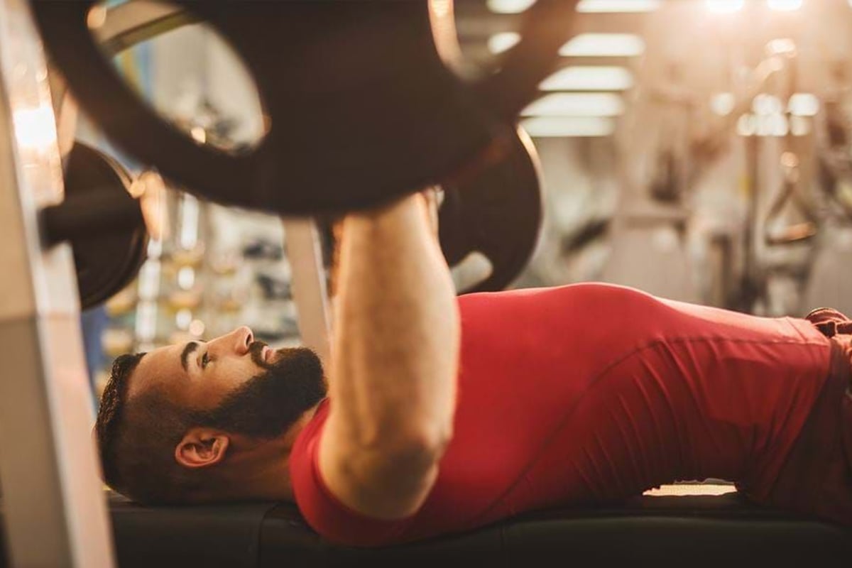 Man in red shirt lifting barbell on bench press in gym with sunlight glare