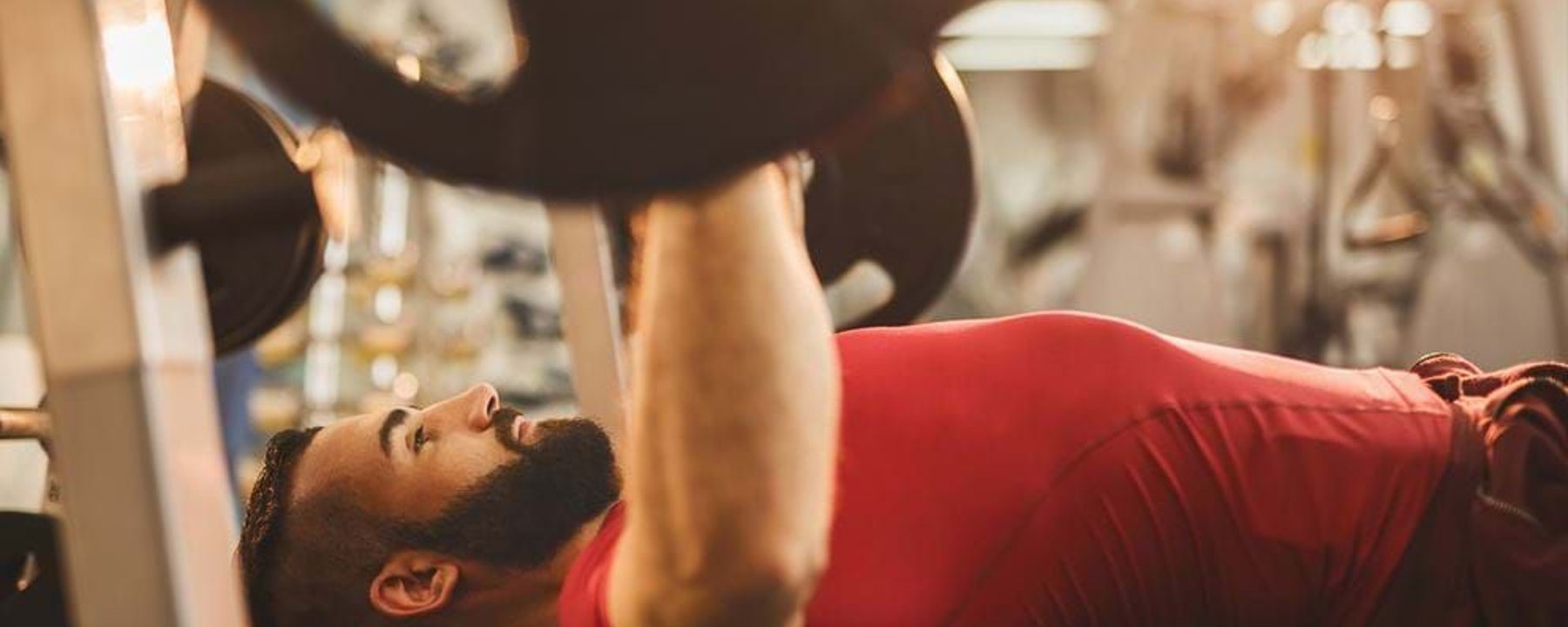 Man in red shirt lifting barbell on bench press in gym with sunlight glare