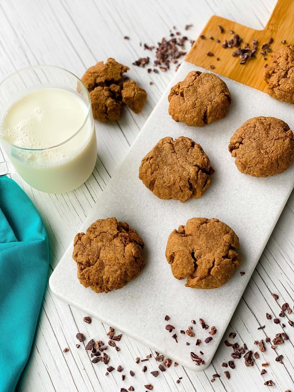 Six brown cookies on a white board with a glass of milk and scattered cocoa nibs nearby