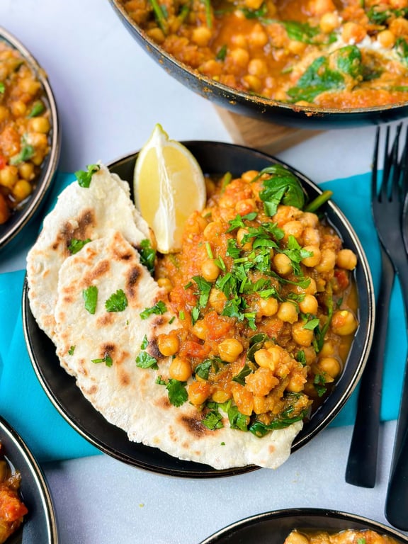 Chickpea curry with spinach, naan bread, and a lemon wedge on a black plate.