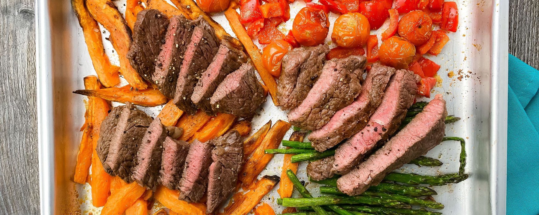 Sliced steak with roasted peppers, tomatoes, sweet potato and asparagus on a baking tray