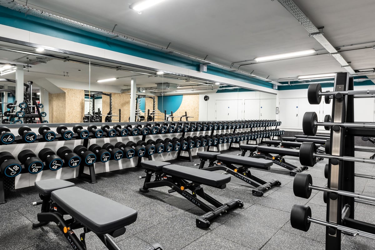 PureGym Manchester First Street: Rows of dumbbell racks, weight benches, barbells and weight plates on rubberized flooring.